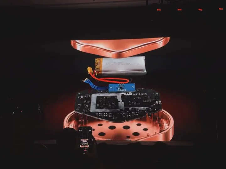 An exploded view of Curapod-M on a big screen: the rose-gold top shell, the battery, the circuit board and the LED base, with an audience phone filming it.